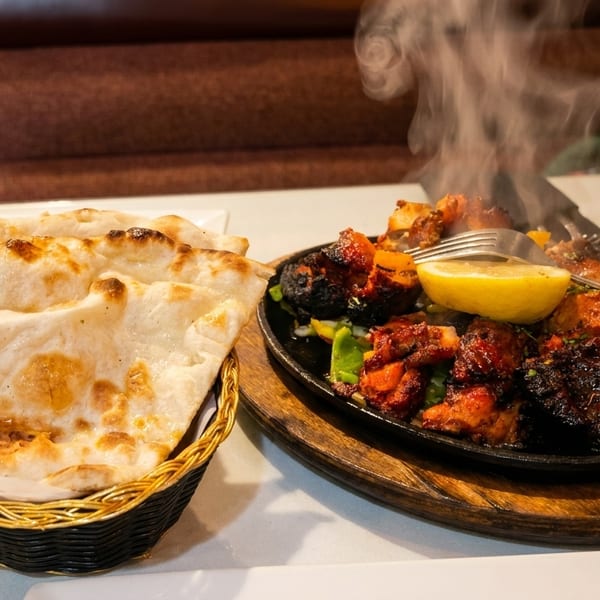 Steaming hot chicken tikka sizzler with a lemon wedge and a basket of naan bread.