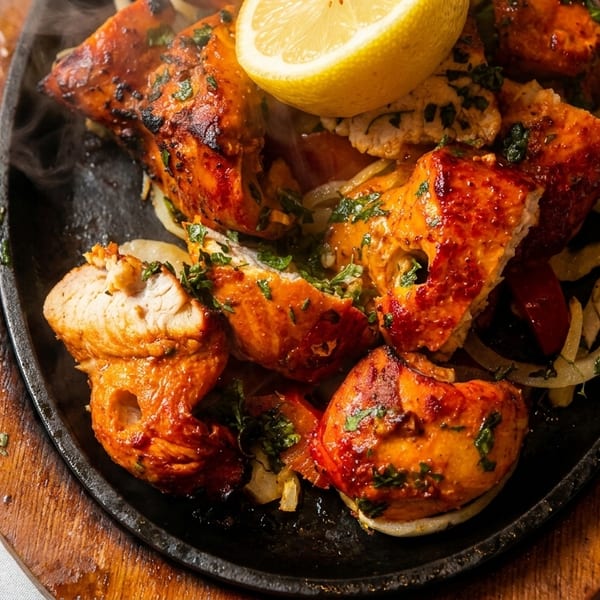Sizzling tandoori chicken with onions and a lemon wedge on a cast iron platter.