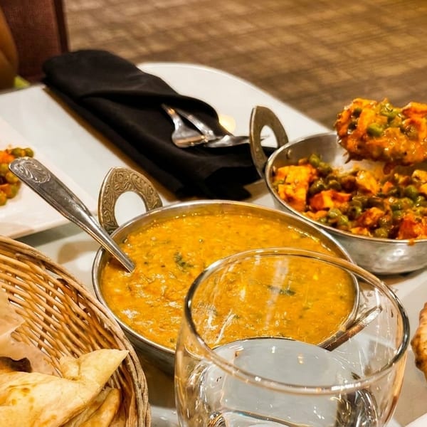 A person spoons Indian curry from a metal bowl, with naan bread and other dishes on a table.