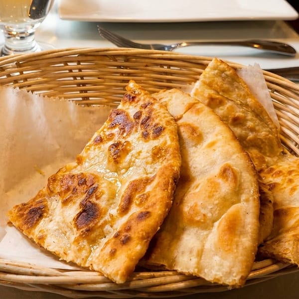 Golden-brown naan bread pieces, some with charred spots, served in a woven basket lined with parchment paper.